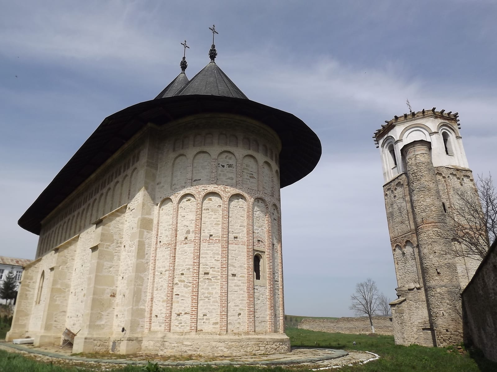 Biserica de piatră a Mănăstirii Dobrovăț, cu turlă neagră și brâuri de cărămidă, lângă turnul-clopotniță de piatră