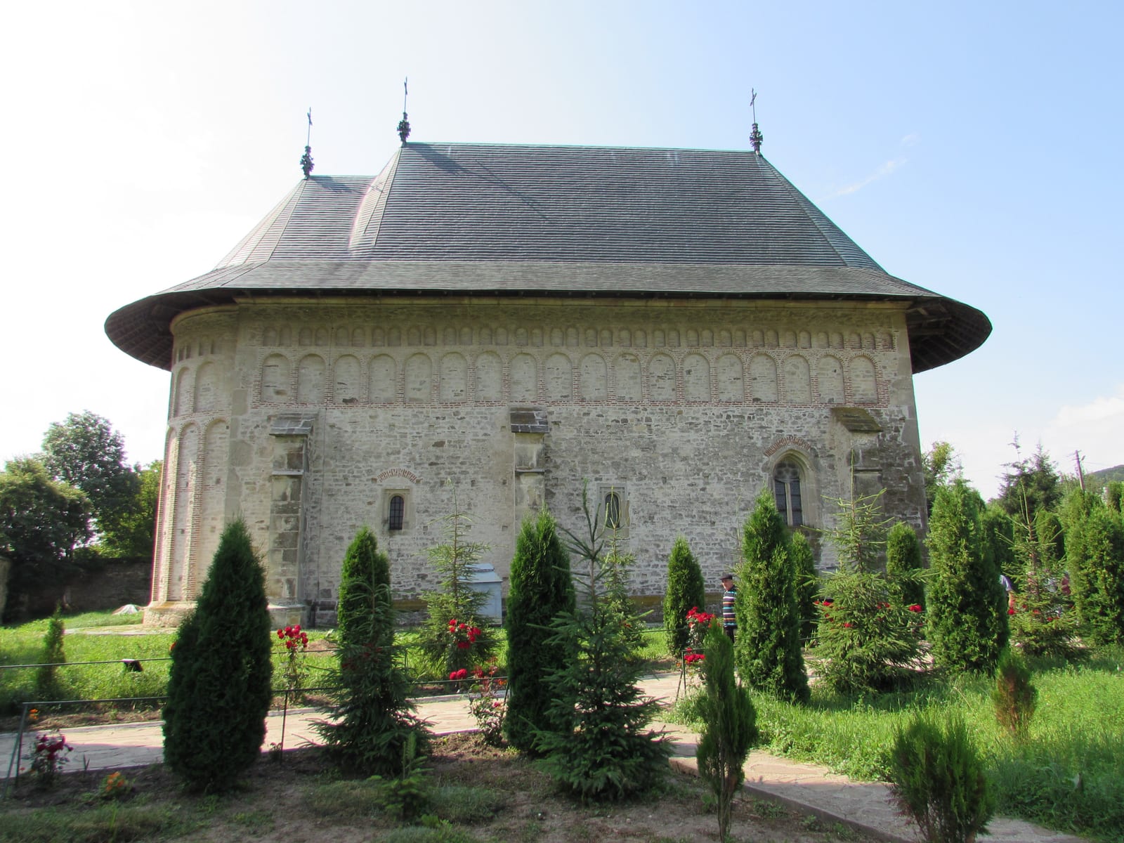Biserica Mănăstirii Dobrovăț văzută din față vara, cu grădina de tuia și trandafiri roșii în curte