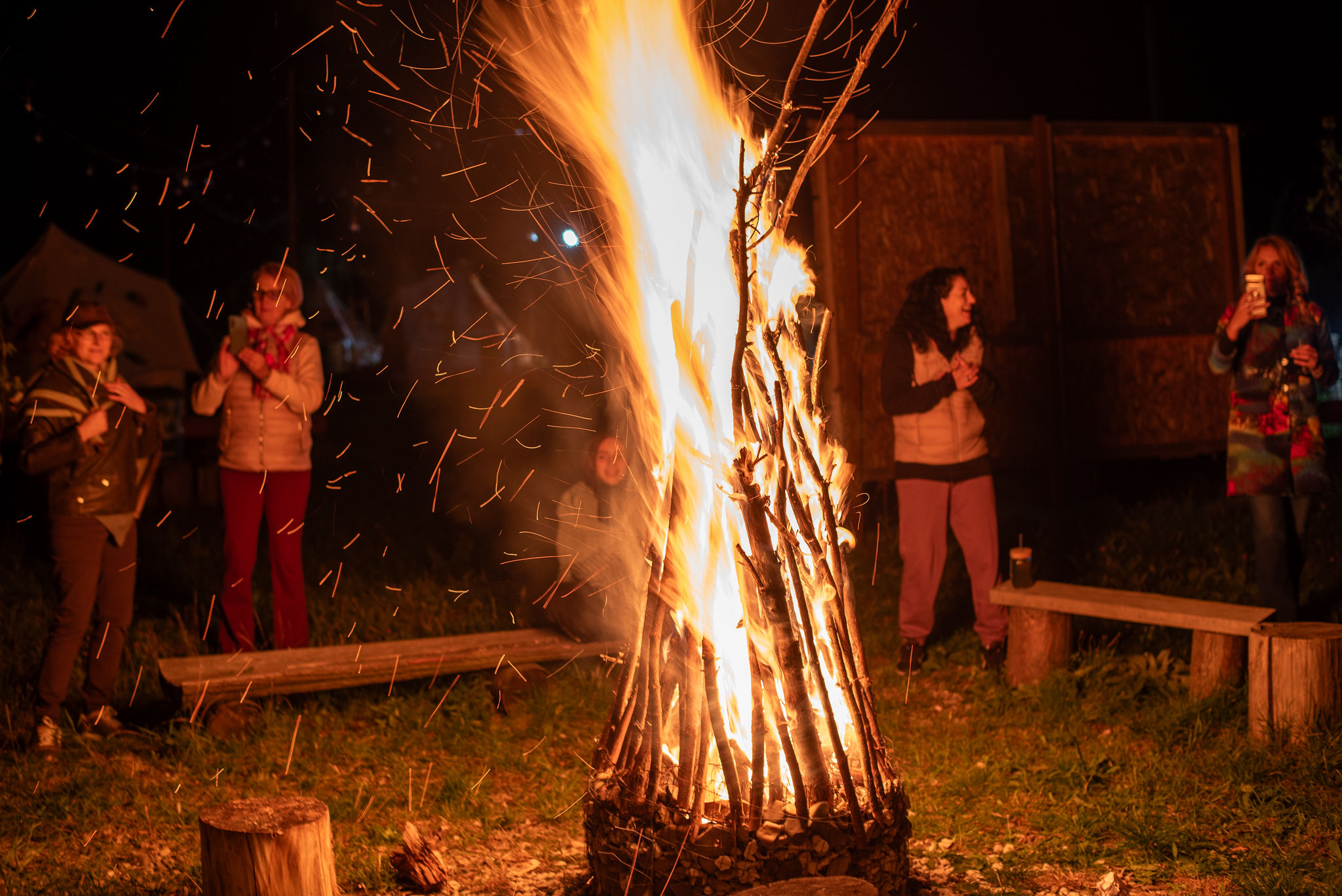 Foc de tabără seara — vâlvătaie cu scântei deasupra cercului de oaspeți